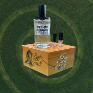 May include: A clear glass perfume bottle with a black cap and the text "STEAMED RAINBOW" is displayed on a decorative wooden box. Two small vials and a perfume sample are also on the box. The box has floral designs.