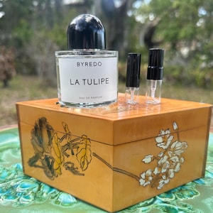 May include: A perfume bottle labeled "BYREDO LA TULIPE EAU DE PARFUM" with a black cap sits atop a decorative wooden box. Two small perfume vials are next to the bottle. The box is a light brown color with floral designs.