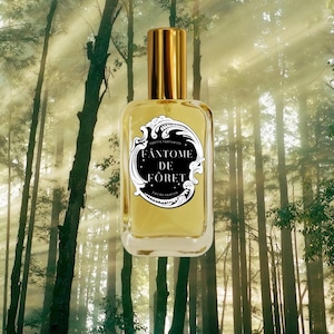 May include: A rectangular glass perfume bottle filled with a golden liquid, featuring a gold cap and a black oval label with white ornate borders. The label displays the text Odette Parfum Co. Fantome De Foret Eau De Parfum in white.