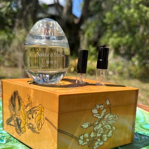 May include: A clear, oval-shaped perfume bottle labeled "Sakura Snow" sits atop a decorative wooden box. The box is painted with floral designs in yellow and white. Two small vials with black caps are also present.
