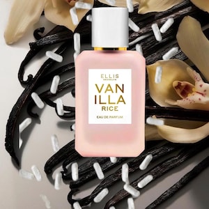 May include: A rectangular, frosted light pink glass perfume bottle with a white cap and a white label featuring the gold text ELLIS BROOKLYN VANILLA RICE EAU DE PARFUM. The bottle is surrounded by dark brown vanilla beans, cream-colored flowers, and white rice grains.