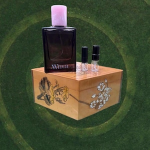 May include: A dark perfume bottle with a pink cap and the word "Witch" on the front, sits on a decorative wooden box. Two small vials are next to the bottle. The box is decorated with floral designs.
