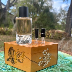 May include: A clear glass perfume bottle with a black cap labeled "NO 1" sits atop a wooden box with floral designs. Two small sample vials are next to the bottle. The box is on a decorative green and brown surface.