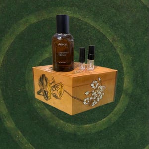 May include: A brown glass bottle of Aesop Marrakech Intense Eau de Parfum, with two small vials, sits atop a wooden box with floral designs. The box is a light brown color.