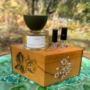 May include: A perfume bottle with a dark green cap and a label that reads "I-Dream MAISON J'ETTO" sits atop a decorative wooden box. Two small sample vials are next to the perfume bottle. The box is decorated with floral designs.