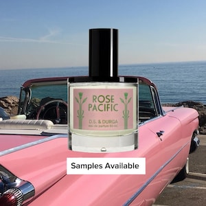 May include: A clear glass perfume bottle with a black cylindrical cap. The label is pink with green floral illustrations and the text: ROSE PACIFIC, D.S. & DURGA, eau de parfum 50 mL. Below the bottle, a white rectangular sign reads: Samples Available.