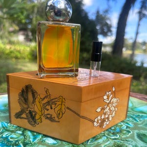 May include: A perfume bottle with a silver cap and a clear, rectangular glass body filled with yellow and green liquid. It sits atop a wooden box with floral designs. A small sample vial is next to the bottle. The box is on a decorative plate.