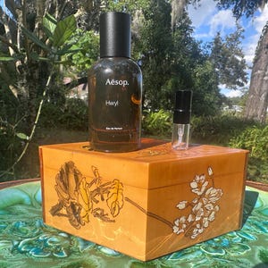 May include: A brown glass bottle of Aesop Hwyl Eau de Parfum and a small sample bottle sit atop a wooden box with floral designs. The box is a warm, golden color, and the background features greenery and a body of water.