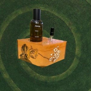 May include: A brown glass bottle of Aesop Virēre Eau de Parfum and a small sample vial sit atop a decorative wooden box. The box is a warm, light brown with floral and leaf designs. The background is a dark green, textured surface.