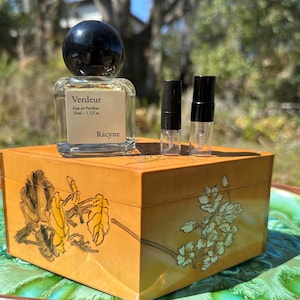 May include: A perfume bottle with a black cap and a label that reads "Verdeur Eau de Parfum 50ml - 1.7 fl oz" sits atop a decorative wooden box. Two small perfume vials are next to the bottle. The box has floral designs.