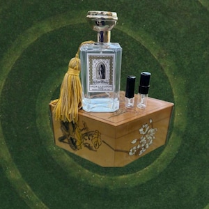 May include: A clear perfume bottle with a silver cap and a yellow tassel sits on a wooden box. The bottle has a label with text and a small illustration. Two small sample vials are next to the bottle.