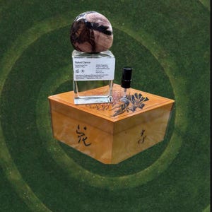 May include: A perfume bottle with a decorative stone top, labeled "Naked Dance," sits atop a wooden box with floral and script details. A small sample vial is next to the bottle. The background is a green, circular pattern.