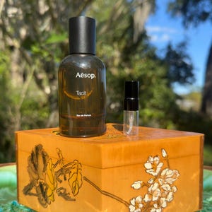 May include: A brown glass bottle of Aesop Tacit Eau de Parfum with a black cap, and a small sample bottle, sit atop a decorative wooden box. The box is painted with floral and leaf designs in shades of yellow and brown. The background is blurred with green foliage.