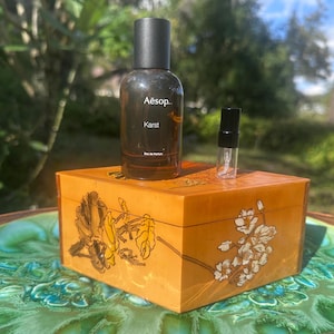 May include: A brown glass bottle of Aesop Karst Eau de Parfum and a small sample bottle sit atop a wooden box with floral designs. The box is placed on a decorative green and blue plate, with a natural background.