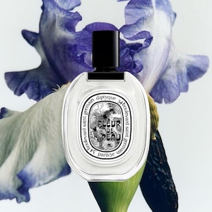 May include: A clear glass perfume bottle with a black cylindrical cap and an oval label featuring the text Diptyque 34 boulevard saint germain Fleur de Peau Paris 5e, positioned in front of a large, light purple and white iris flower bloom.