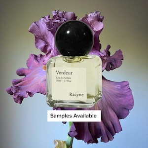 May include: A clear glass perfume bottle with a black spherical cap, labeled "Verdeur" and "Racync", sits in front of a large purple iris flower. The bottle contains 1.7 fl oz of perfume. The text "Samples Available" is at the bottom.