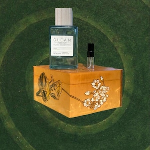 May include: A rectangular turquoise perfume bottle with a wooden cap and a small sample bottle sit atop a decorative wooden box. The box is adorned with floral artwork in yellow and white against a warm orange background. The background is a dark green.
