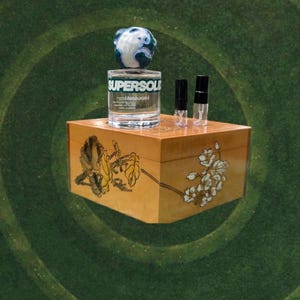 May include: A clear glass perfume bottle with the text "SUPERSOL" and a decorative globe-shaped top. The bottle sits on a wooden box with floral artwork. Two small vials are also on the box.