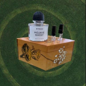 May include: A clear glass perfume bottle with a black cap and the text "BYREDO MOJAVE GHOST" sits atop a decorative wooden box. Two small perfume vials with black caps are next to the bottle. The box has floral designs.