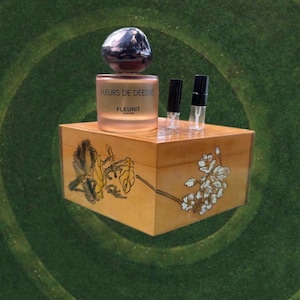 May include: A perfume bottle with a silver cap and the text "FLEURS DE DÉESSE FLEURIT" sits atop a decorative wooden box with floral designs. Two small vials are next to the perfume bottle. The background is a green, circular pattern.