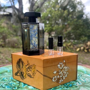 May include: A dark perfume bottle with a black cap and a label with blue and white details sits atop a wooden box with floral designs. Two small sample vials are next to the bottle. The box is on a decorative surface.