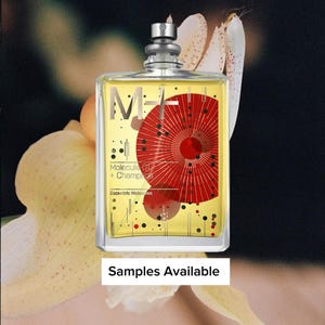 May include: A rectangular perfume bottle with a silver cap and a yellow label featuring red and black abstract designs. The text "Molecule 01 + Champaca" is visible. The background includes a blurred floral arrangement. Text on the image reads "Samples Available."