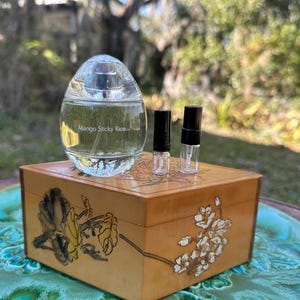 May include: A clear, oval-shaped perfume bottle with the text "Mango Sticky Rice" sits atop a wooden box with floral artwork. Two small sample vials with black caps are also on the box. The box is on a decorative, turquoise-colored surface.