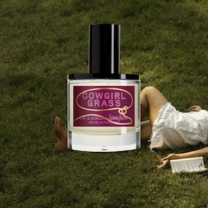 May include: A clear glass perfume bottle with a black cylindrical cap and a rectangular label. The label features a maroon background with the text COWGIRL GRASS, D.S. & DURGA, Neiman Marcus, and eau de parfum 50 ml printed in white and gold.