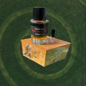 May include: A perfume bottle with a black cap and a label that reads "Musc Ravageur" sits on a decorative wooden box. A small sample vial is next to the bottle. The box has a floral design.