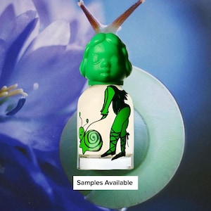 May include: A perfume bottle with a green cap shaped like a face. The bottle has a white base with a green illustration of a person and a snail. The text "Samples Available" is at the bottom of the image. The background is a blue and purple floral design.