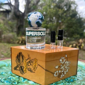 May include: A clear glass bottle with the word "SUPERSOL" in green lettering, topped with a globe-shaped stopper. Two small vials with black caps sit beside the bottle on a wooden box with floral designs.