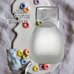 May include: A frosted glass perfume bottle with a silver cap featuring a heart logo and the text LORE LIL SOFTIE SAMPLE. The bottle is placed over a white palette decorated with colorful ring-shaped cereal pieces, a small white kitten illustration, and a metal tool.