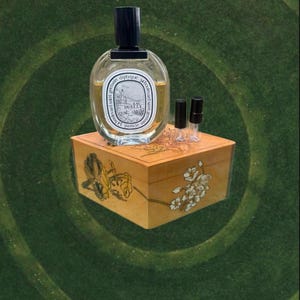 May include: An oval perfume bottle with a black cap sits atop a decorative wooden box. The box is adorned with floral designs. Two small vials with black caps are next to the perfume bottle. The background is a green, swirled pattern.