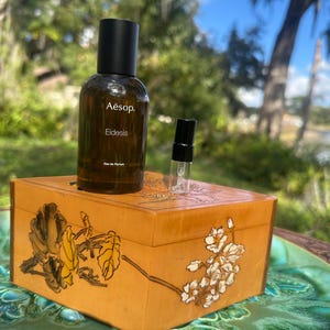 May include: A brown glass perfume bottle with a black cap and the text "Aesop. Eidesis" sits on a wooden box with floral designs. A small sample vial is next to the bottle. The box is on a green ceramic plate.