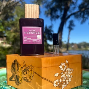 May include: A dark purple perfume bottle with a wooden cap and a small sample bottle sit on a decorative wooden box. The perfume bottle has a label that reads "Clean Reserve". The box is decorated with floral designs.