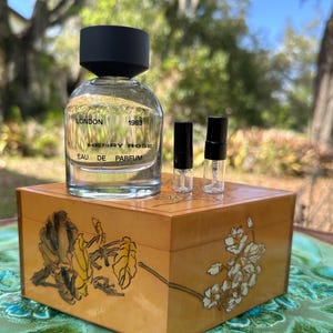 May include: A clear glass perfume bottle with a black cap and the text "LONDON 1983 HENRY ROSE EAU DE PARFUM" sits on a wooden box with floral designs. Two small perfume vials are also on the box. The box is on a green and blue decorative plate.