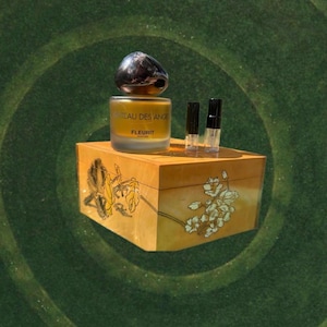 May include: A perfume bottle with a dark, rounded cap sits atop a wooden box with floral and leaf designs. The bottle is labeled "CHATEAU DES ANGE FLEURIT PERFUMES." Two small vials are next to the bottle. The box is on a green background.