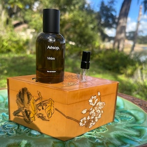 May include: A dark brown perfume bottle with a black cap and the text "Aesop. Virère" sits on a wooden box with floral designs. A small sample vial is next to the bottle. The box is on a green and brown decorative table.