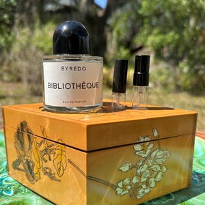 May include: A perfume bottle with a black cap and the text "BYREDO BIBLIOTHÈQUE EAU DE PARFUM" sits atop a decorative wooden box. Two small perfume vials are next to the bottle. The box is yellow with floral designs.