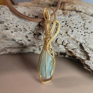 May include: A gold-colored wire-wrapped pendant with a light blue gemstone. The pendant is suspended from a brown leather cord. The gemstone is teardrop-shaped. The wire is intricately woven around the stone, creating a cage-like design.