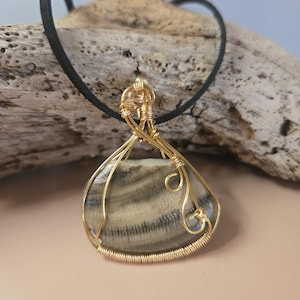 May include: A handcrafted pendant necklace featuring a teardrop-shaped stone with gray and beige stripes, encased in gold-colored wire. The pendant hangs from a black cord necklace. The background is a light pink surface and a piece of driftwood.