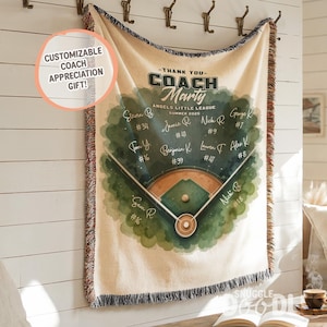 May include: A beige baseball-themed throw blanket with a watercolor design. The blanket features a baseball field graphic and the text "Thank You Coach Marty" with player names and numbers. The blanket has a fringe edge and is hanging on a wall.