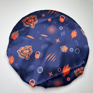 Puede incluir: Un gorro de satén azul marino con un patrón repetitivo de logotipos de cabezas de oso naranjas, huellas de oso, pinceladas naranjas y pequeños círculos y cruces blancas dispersos por la superficie circular de tela suave.