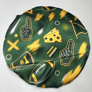Puede incluir: Un gorro de satén verde oscuro con un patrón repetitivo de dedos de espuma amarillos, un trozo de queso suizo, un balón de fútbol americano, un megáfono con el texto #1 y pequeñas banderas, acentuado por texturas de pinceladas amarillas y blancas.