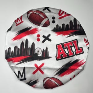 Puede incluir: Un gorro de satén con fondo blanco y un patrón repetitivo de balones de fútbol americano rojos, siluetas de ciudades negras, coronas rojas, marcas X negras y el texto ATL en rojo y negro, con trazos de pincel abstractos.