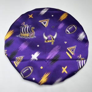 Puede incluir: Un gorro de satén morado con un patrón repetitivo de barcos vikingos, cascos con cuernos y balones de fútbol americano, acentuado con diseños de pinceladas abstractas en amarillo, blanco y negro, junto con pequeñas formas geométricas.