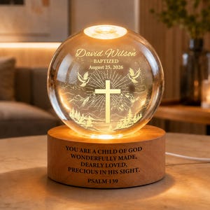 May include: A clear glass sphere lamp on a cylindrical wooden base. The sphere features an etched cross, two doves, mountains, trees, and the text: David Wilson BAPTIZED August 25, 2026. The base displays: YOU ARE A CHILD OF GOD PSALM 139.