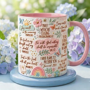 May include: A white ceramic mug with a pink interior and handle, featuring embossed floral, butterfly, rainbow, and globe motifs alongside various Bible verses including Psalm 30:5, 1 Thessalonians 5:17, Luke 1:37, Jeremiah 29:11, and 2 Corinthians 5:7.