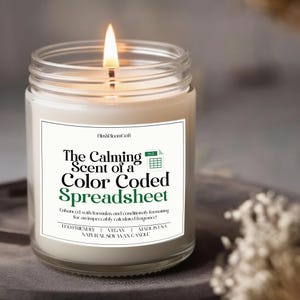 May include: A lit soy wax candle in a clear glass jar. The label reads "The Calming Scent of a Color Coded Spreadsheet." The candle is eco-friendly, vegan, and made in the USA. The candle has a white wax and a silver wick.