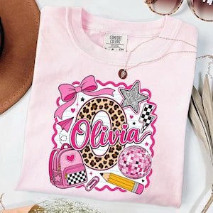 Pode incluir: Uma t-shirt rosa claro com um design gráfico que apresenta o nome Olivia em letra cursiva rosa, uma letra O grande com padrão de leopardo, um laço rosa, uma mochila rosa, uma bola de discoteca, um lápis amarelo, uma estrela prateada e um raio preto e branco.